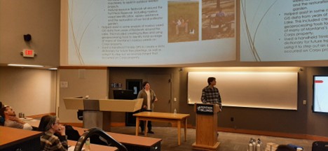 large auditorium with presenters in front of two large screens