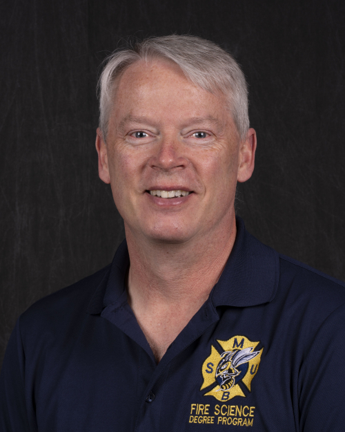 A smiling, clean-shaven man with gray hair wearing a firefighter shirt.