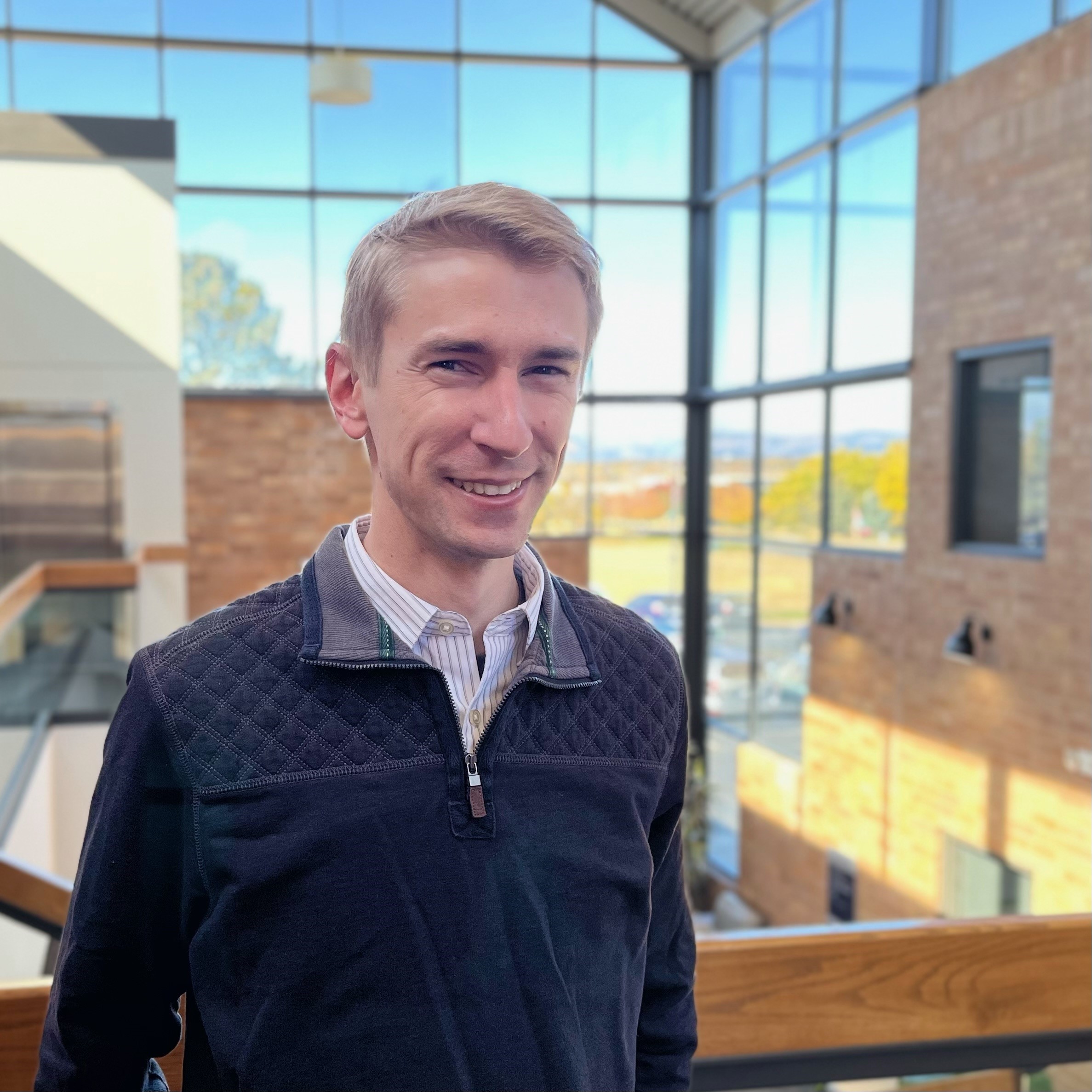A smiling man with combed blond hair wearing a blue sweater and standing in a glass atrium.