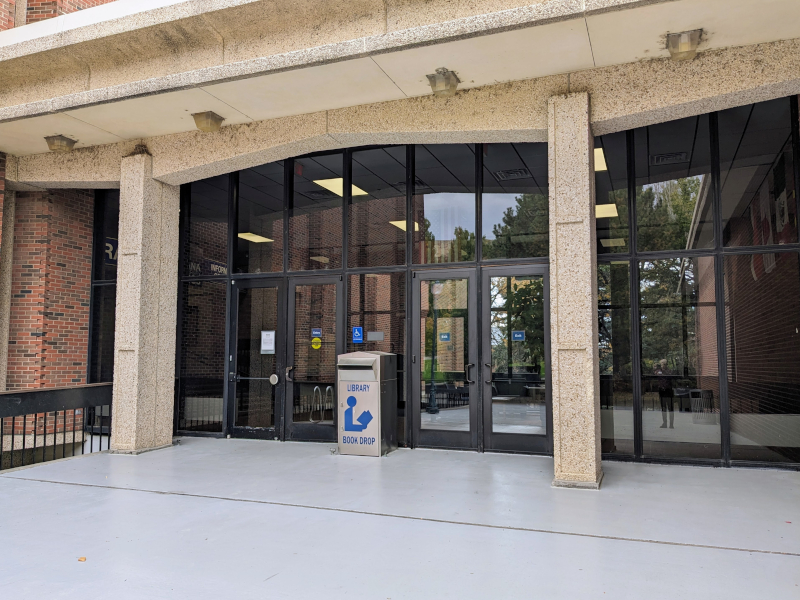 A set of doors set into a glass wall. A silver book return bin stands in front.