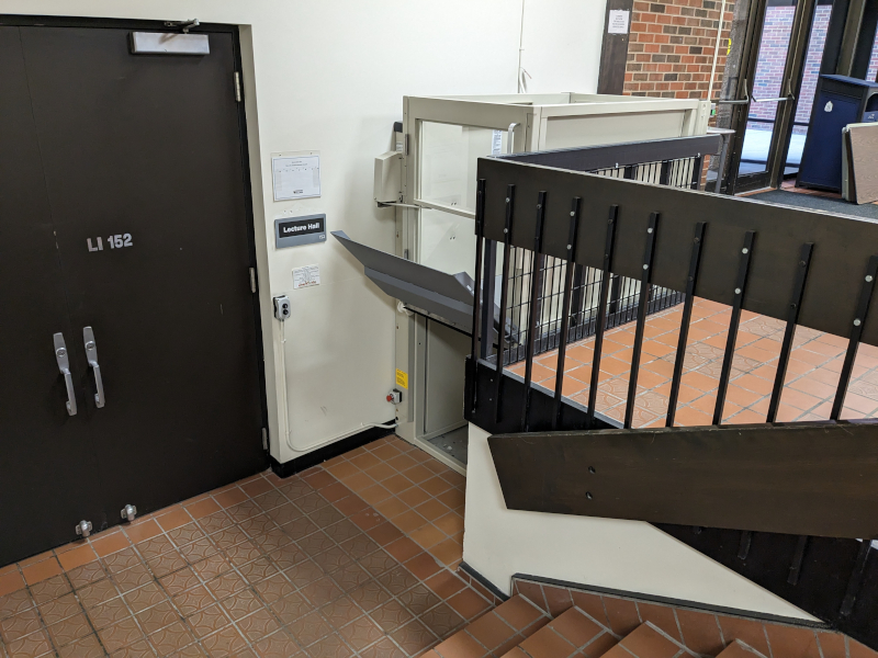 A lift in a lobby, bypassing a half-flight of stairs to get to a door marked "Lecture Hall."