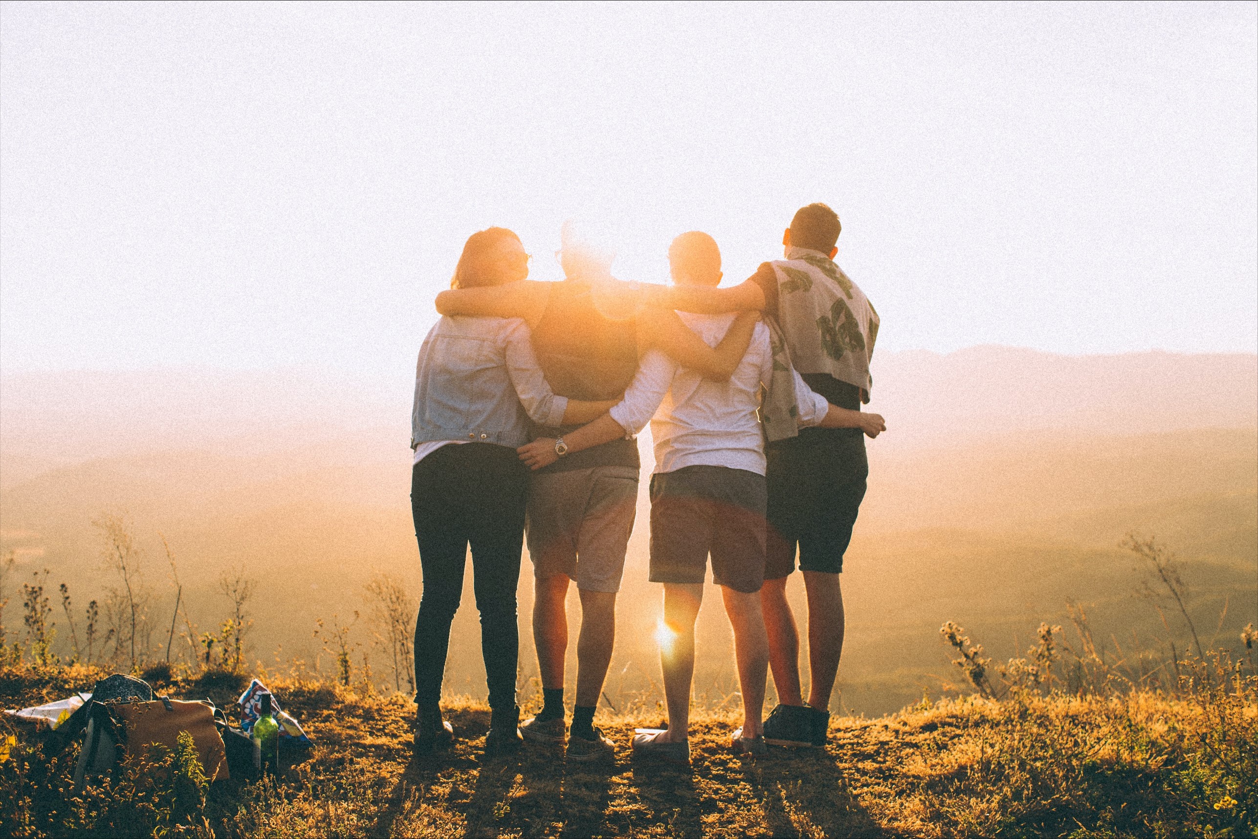 Four people stand with their backs to the camera and arms around each others shoulders. They are looking toward the sun, which shines toward to viewer. 