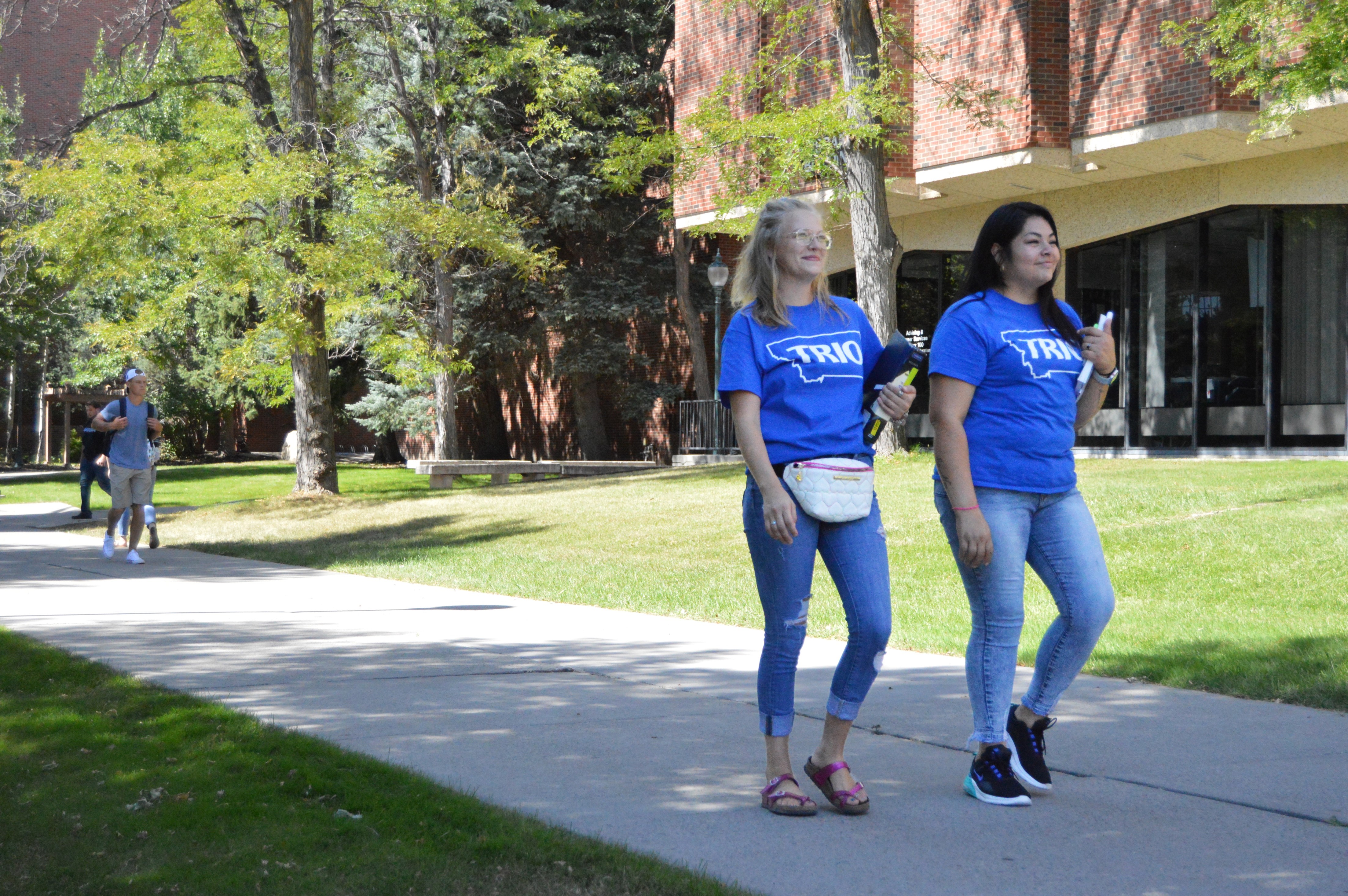 Two students walk in front of the MSUB library. They are both wearing blue TRIO t-shirts and smiling.